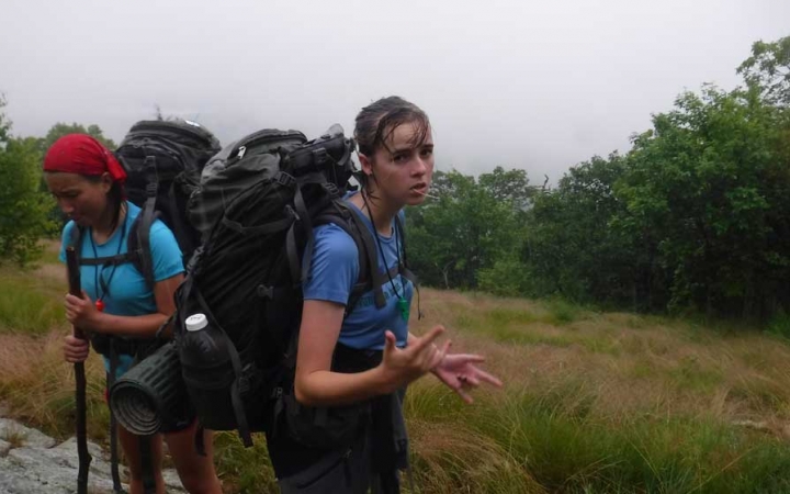 Two people wearing backpacks stand in a grassy meadow surrounded by trees. One of them gives an "I love you" hand gesture to the camera. 
